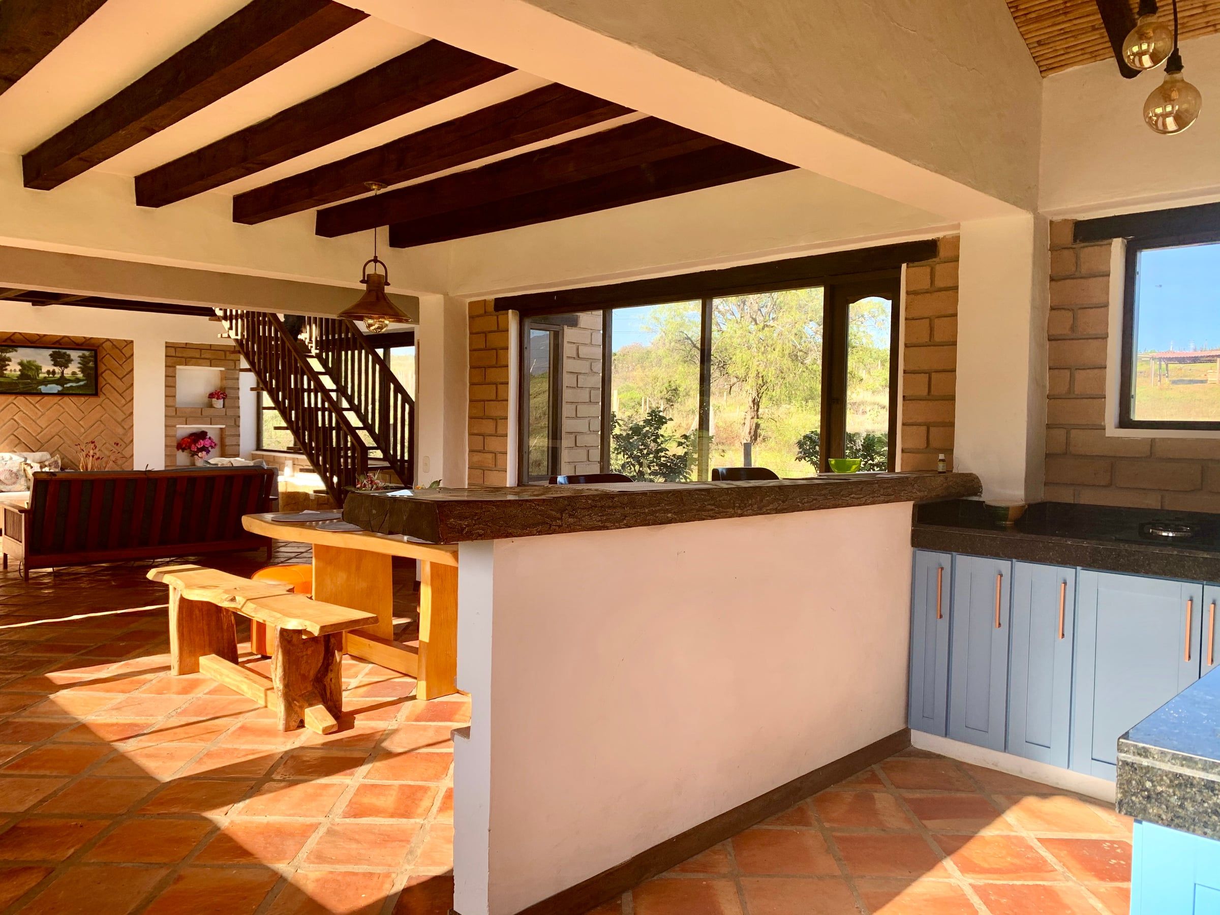 Cocina abierta con barra, escalera de madera al fondo y piso de baldosa de barro bañado por luz natural