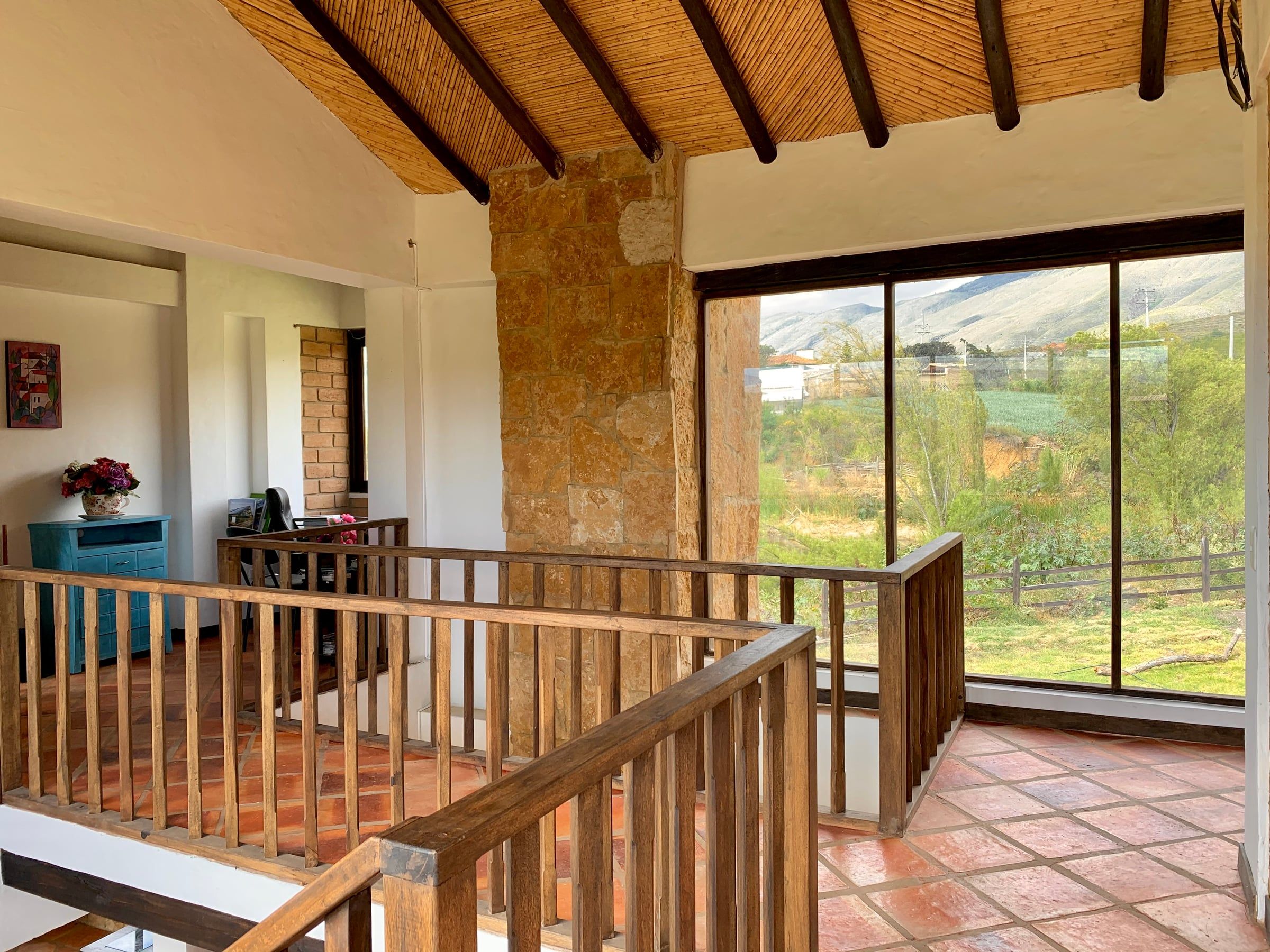 Hall del segundo piso con baranda de madera, muro de piedra y ventanal hacia el paisaje