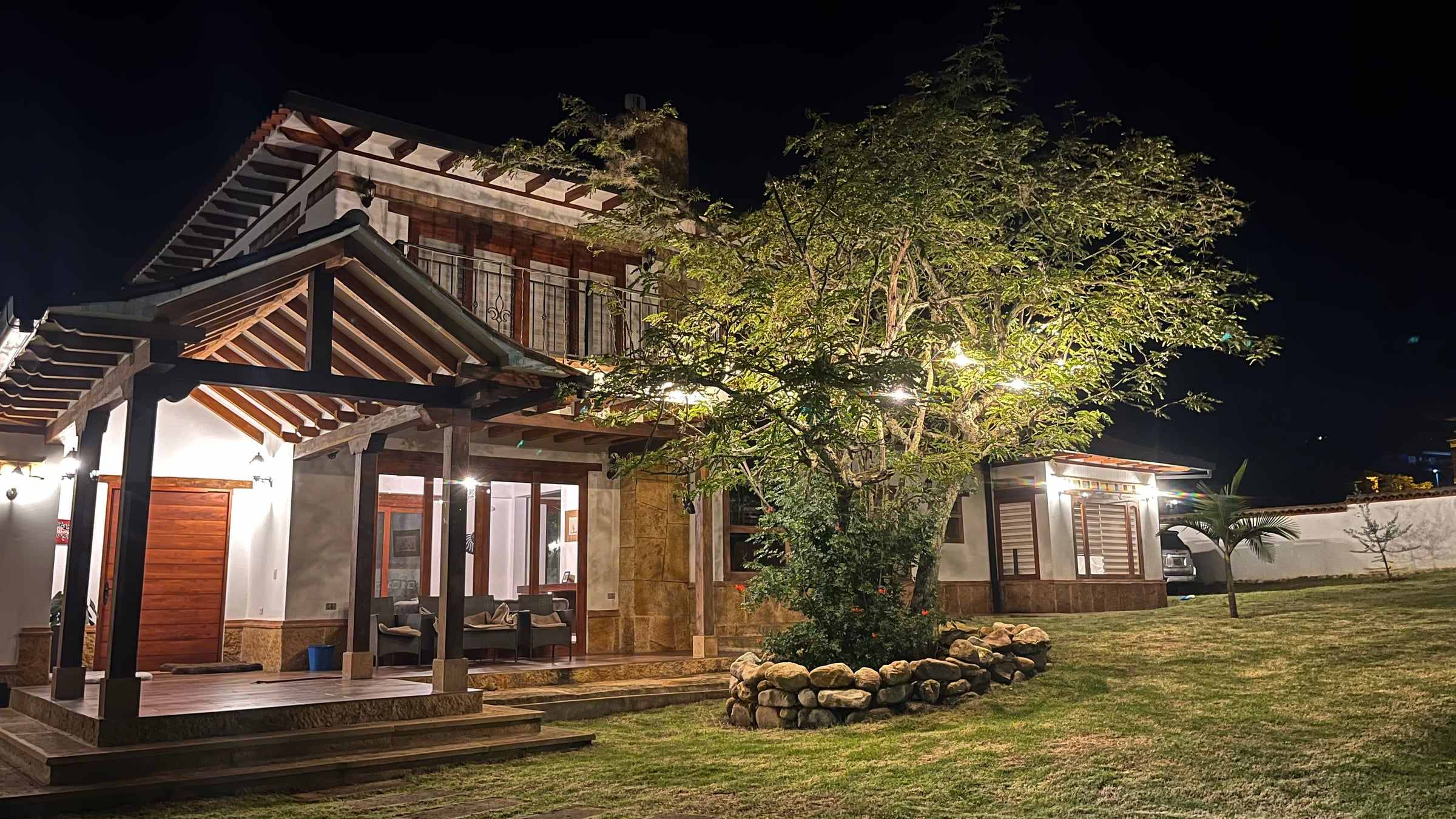 Fachada nocturna iluminada de la Casa Dividivi, con corredor de madera, cubierta de teja de barro y un árbol en el jardín, en la vereda El Roble de Villa de Leyva