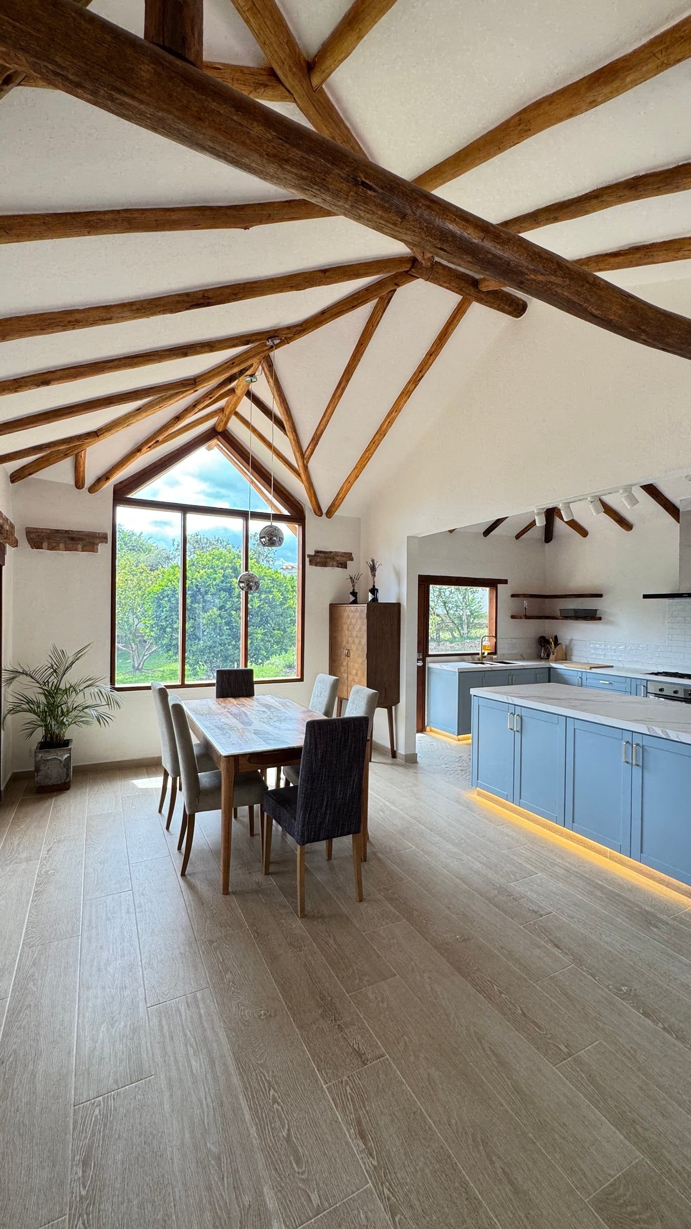 Casa Nido Verde después de la remodelación: sala-comedor con piso claro tipo madera, mesa de comedor, ventana en punta y cubierta de vigas de madera conservada